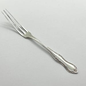 May include: A silver-toned metal cocktail fork featuring three slender tines and a decorative handle with a raised beaded border design. The handle tapers towards the neck and widens at the base, showcasing a polished finish and intricate detailing.
