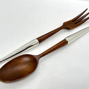 May include: A wooden fork and spoon set with silver-coloured metal accents. The fork has three tines, and the spoon has a rounded bowl. The handles are a dark brown colour, and the metal accents are silver. The set is designed for serving.