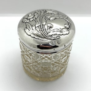 May include: A silver-toned jar with a domed lid featuring a floral design. The clear glass jar has a diamond-cut pattern. The jar is cylindrical and sits on a white surface. The lid is highly reflective.