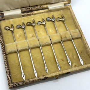 May include: A set of six silver cocktail picks, each featuring a detailed rooster finial at the top. The slender metal picks have pointed ends and are arranged in a rectangular presentation case with a textured exterior and a soft, velvet-lined interior.