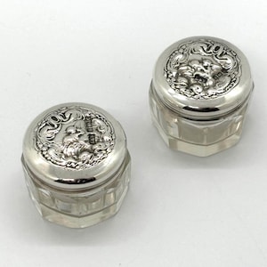 May include: Two antique silver-topped glass jars. The lids feature an ornate relief design. The jars have a faceted, clear glass body. These jars are likely for storing small items or cosmetics.
