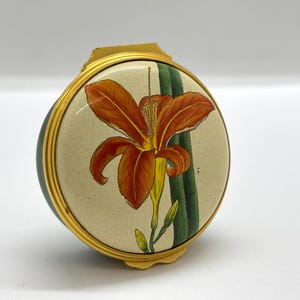 Peut inclure: Un poudrier rond à bordure dorée avec un couvercle de couleur crème orné d'une illustration détaillée d'un lys orange avec des tiges vertes. Le boîtier en métal présente une finition dorée polie et une fermeture à charnière sur le dessus.