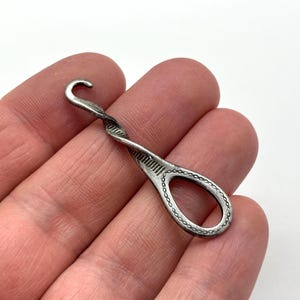 以下が含まれることがあります： 螺旋状にねじれたハンドルと、ベースに涙型のループを備えた、銀色の金属製ボタンフック。ハンドルには質感のあるリブデザインが施されており、ループには繊細なチェーンリンク模様がエンボス加工されています。