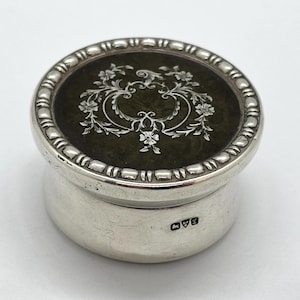 May include: A small, round silver trinket box. The lid is dark green with a white floral design. The box has a beaded edge and a shiny surface. The base has a hallmark. The box is approximately 5 cm in diameter.