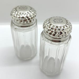 May include: Two clear glass shakers with vertical fluted sides and removable silver-toned metal caps. The caps feature a hammered texture and a circular top plate engraved with a decorative monogram consisting of the intertwined letters A, H, and C.