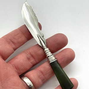 May include: A small silver butter knife featuring a scalloped edge blade and a dark green handle. The handle is connected to the blade by a silver ferrule with horizontal ribbed detailing. The knife has a polished metallic finish.