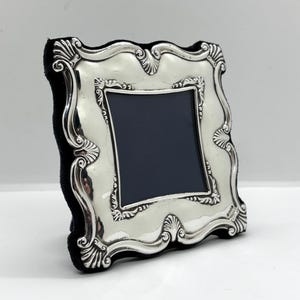 May include: A silver picture frame with an elaborate, scalloped design. The frame has a black backing and a dark blue interior. The frame is made of a shiny, reflective material and has a classic, elegant style.