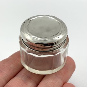 May include: A small, clear glass jar with a faceted, multi-sided body and a polished, reflective silver-toned metal lid. The lid features a slightly wavy, organic edge design, contrasting with the structured, geometric shape of the glass base.