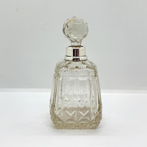 May include: A clear, vintage perfume bottle with a faceted crystal stopper and silver-tone collar. The bottle has a square base with diamond-shaped cut glass details. The stopper is a large, clear, faceted crystal ball.