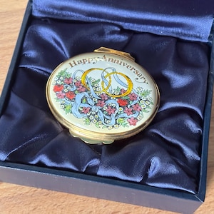 May include: A round gold-toned metal trinket box with a hinged lid featuring the text Happy Anniversary, two interlocking gold rings, and a colorful floral arrangement with blue ribbons, presented inside a dark blue satin-lined gift box.