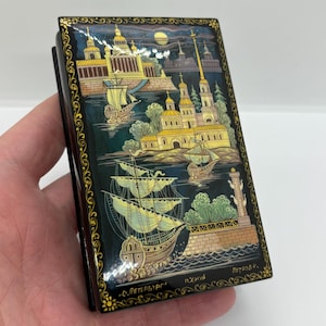 May include: A rectangular, black lacquered box with a detailed painting of a cityscape with ships, buildings, and a lighthouse. The artwork features gold accents and the text "С.Петербург" and "Петров".