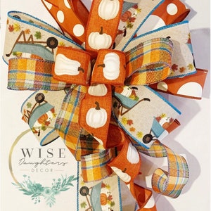 May include: A fall-themed bow made with various ribbons. The bow features a plaid ribbon, a ribbon with a white pumpkin pattern, a ribbon with a white polka dot pattern, and a ribbon with a wheelbarrow and pumpkin pattern. The bow is tied with a blue ribbon.