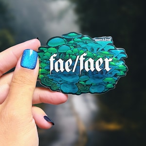 May include: A clear sticker featuring a cluster of blue and green mushrooms with the text "fae/faer" in white. The sticker has a dark background and is held by a hand with blue glitter nail polish.
