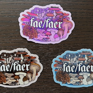 May include: Three stickers featuring a cluster of colourful mushrooms with the text "fae/faer" in a gothic font. The stickers are on a wooden surface. The stickers are in shades of purple, white, and blue.