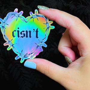 Puede incluir: Una pegatina holográfica con forma de corazón arcoíris rodeada de alambre de púas. El texto "cisn't" está escrito en fuente gótica negra dentro del corazón.