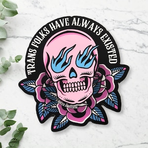 May include: Pink skull with blue flames for eyes and eyelashes, surrounded by pink and blue roses with green leaves. The text "TRANS FOLKS HAVE ALWAYS EXISTED" is in a black circle around the skull.