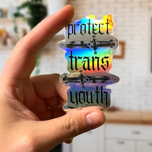 Puede incluir: Pegatina holográfica con el texto "protect trans youth" en fuente gótica, con dos ilustraciones de dagas. La pegatina tiene un brillo arcoíris y se sostiene en una mano.