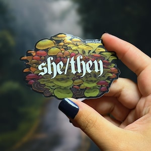 Puede incluir: Una pegatina con un diseño de varios hongos en tonos verdes, amarillos, naranjas y rojos. Las palabras "she/they" están en blanco y en negrita. También está presente el texto "queer & feral".