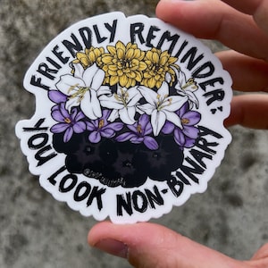 Può includere: Un adesivo bianco con un bordo nero e il testo "Friendly Reminder: You Look Non-Binary" in un design circolare. Il testo è circondato da un bouquet di fiori gialli, bianchi e viola.