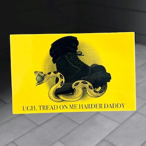 May include: A yellow rectangle featuring a black boot stepping on a yellow and tan snake. The text "UGH, TREAD ON ME HARDER DADDY" is at the bottom. The words "@queer.and feral" are on the boot.