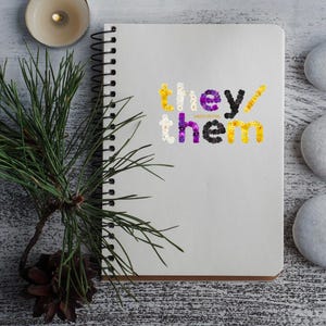 May include: A white notebook with a spiral binding. The words "they/them" are spelled out in yellow, purple, and black flowers on the front cover.