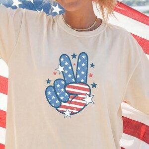 May include: Ivory-colored t-shirt featuring a peace sign design in red, white, and blue, with stars and stripes. The shirt is a relaxed fit. The word "IVORY" is printed at the bottom of the image.