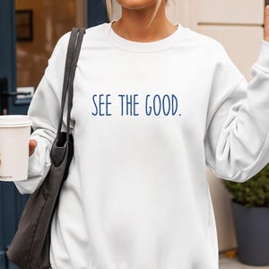 May include: White sweatshirt with the text "SEE THE GOOD" in navy blue. The sweatshirt has a crew neck and long sleeves. The person in the image is holding a coffee cup and a tote bag.