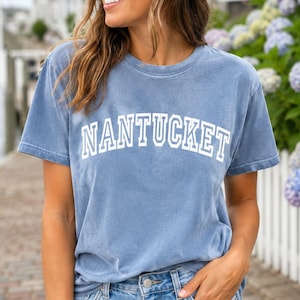 Könnte beinhalten: Ein verwaschenes blaues Kurzarm-T-Shirt mit dem Wort NANTUCKET in weißen Blockbuchstaben auf der Brust. Das Kleidungsstück hat einen Rundhalsausschnitt und eine lockere Passform, gefertigt aus einem weichen, verwaschenen Baumwollstoff.