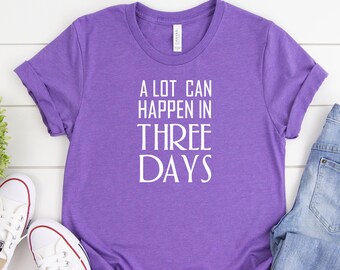 Pode incluir: Uma camiseta roxa com texto branco que diz "A lot can happen in three days". A camiseta está colocada sobre uma superfície de madeira branca com um par de tênis brancos e shorts jeans azuis.