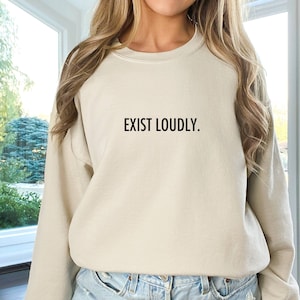 May include: A light beige sweatshirt with the text "EXIST LOUDLY." printed in black on the front. The sweatshirt is being worn by a person wearing denim shorts.