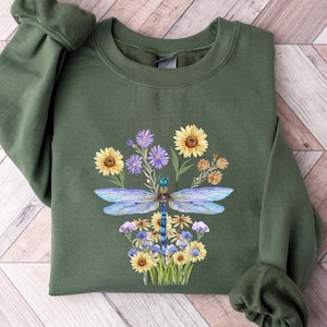 May include: An olive green sweatshirt featuring a detailed illustration of a dragonfly with blue wings, surrounded by sunflowers and purple wildflowers. The design is centered on the front of the long-sleeved top, which is folded on a wooden surface.