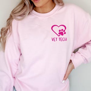 Può includere: Una felpa rosa chiaro con la scritta "VET TECH" sotto un disegno a forma di cuore con un'impronta di zampa all'interno. La felpa è a maniche lunghe ed è realizzata in un materiale morbido.