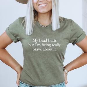 Può includere: Una maglietta verde con il testo "My head hurts but I'm being really brave about it." stampato in bianco.