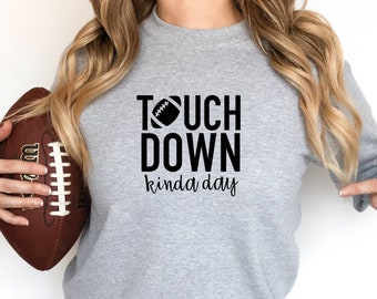 Puede incluir: Una sudadera gris con el texto "TOUCH DOWN kinda day" y un gráfico de fútbol negro. La modelo sostiene un balón de fútbol marrón.