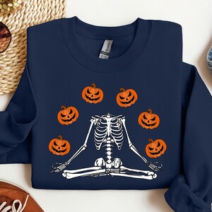 May include: A navy blue sweatshirt featuring a white skeleton meditating, surrounded by orange pumpkins. The skeleton is wearing a white headband.