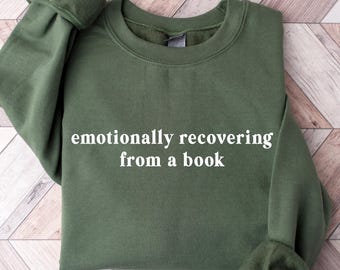 May include: An olive green sweatshirt with the text "emotionally recovering from a book" in white. The sweatshirt is folded on a light brown and white wooden surface. The sleeves are cuffed.