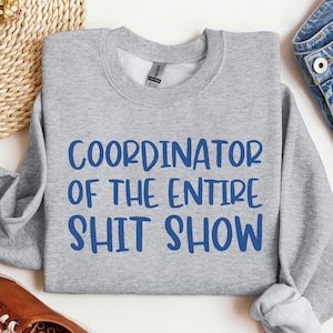 May include: A heather gray sweatshirt with the text "COORDINATOR OF THE ENTIRE SHIT SHOW" in blue. The sweatshirt is displayed with a pair of brown shoes, sunglasses, and a pair of blue jeans.