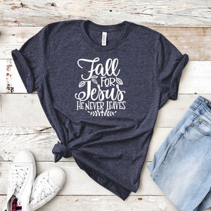 K&ouml;nnte beinhalten: Ein marineblaues T-Shirt mit dem Text "Fall for Jesus He Never Leaves" in wei&szlig;en Buchstaben. Das Shirt liegt flach auf einer wei&szlig;en Holzoberfl&auml;che mit einem Paar wei&szlig;en Sneakers und einer blauen Jeans.