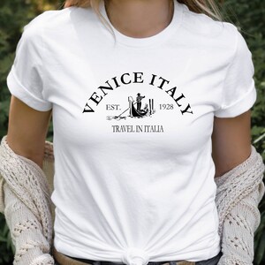 May include: White t-shirt with a black graphic design that says "Venice Italy Est. 1928 Travel in Italia".