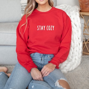以下が含まれることがあります： 白い文字で「STAY COZY」と書かれた赤いスウェットシャツを着た女性。彼女は白いふわふわの敷物の上に座っており、隣には白いニットの毛布がある。