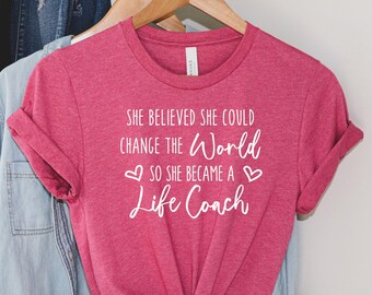 May include: Pink t-shirt with white text that reads "She believed she could change the world so she became a life coach" with two white hearts on either side of the text.