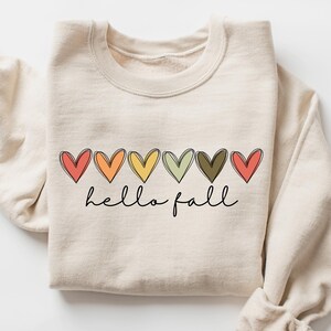 May include: Cream-colored sweatshirt with a row of colorful hearts and the words "hello fall" in a handwritten font. The hearts are in shades of red, orange, yellow, green, and brown. The sleeves are rolled up.