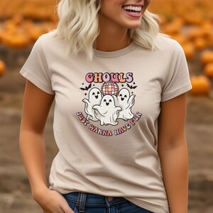 Puede incluir: Una camiseta beige con un gráfico de tres fantasmas bailando alrededor de una bola de discoteca. El texto "Ghouls Just Wanna Have Fun" está escrito en una fuente retro.