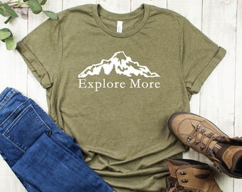 May include: A green t-shirt featuring a white mountain graphic and the text "Explore More". The shirt is styled with blue jeans and brown hiking boots.