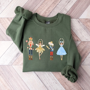 May include: A green sweatshirt with a festive design featuring a nutcracker, a ballerina, a mouse king, and a sugar plum fairy. The characters are all wearing colorful costumes and are dancing.