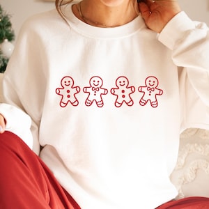Może przedstawiać: Biała bluza z czerwonym nadrukiem przedstawiającym czterech ludzików z piernika. Dwóch ludzików z piernika ma muszki. Bluza ma okrągły dekolt i jest wykonana z miękkiego materiału. Projekt ma świąteczny motyw.