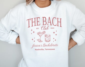 Può includere: Felpa bianca con grafica rossa che recita "THE BACH CLUB EST. 2024" con illustrazioni di cocktail e la scritta "Jessica's Bachelorette Nashville, Tennessee". Il top a maniche lunghe è in stile casual.