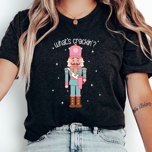May include: A black t-shirt featuring a white graphic of a pink and blue nutcracker holding a cup. The text "What's crackin'?" is above the nutcracker.