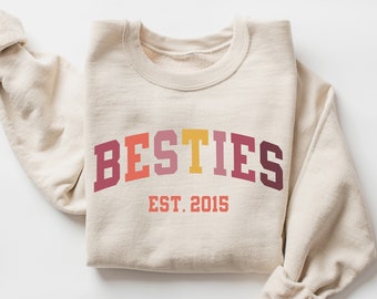 Puede incluir: Una sudadera beige con el texto "BESTIES EST. 2015" en un diseño de arcoíris.