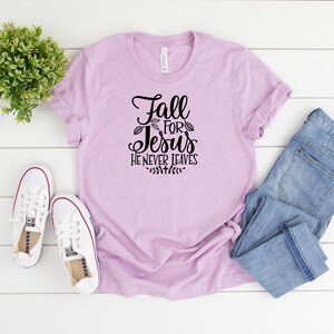 K&ouml;nnte beinhalten: Ein helles lila T-Shirt mit dem Text "Fall for Jesus He Never Leaves" in schwarzer Schrift. Das Shirt liegt flach auf einer wei&szlig;en Holzoberfl&auml;che mit einem Paar wei&szlig;en Turnschuhen und einer blauen Jeans.